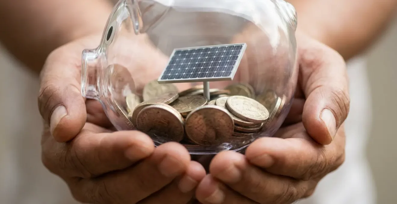 Gros plan de mains tenant délicatement une tirelire transparente contenant des pièces et un panneau solaire miniature